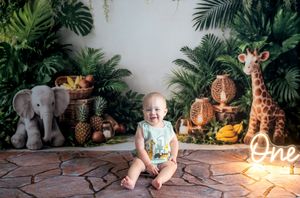 Baby's first birthday cake smash with jungle theme, surrounded by toy animals, giraffe, elephant, and tropical plants.