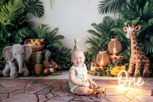 Baby's first birthday jungle safari cake smash with elephant and giraffe stuffed animals, tropical plants, and glowing One sign.