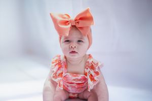Baby wearing a coral pink outfit with ruffles and bow sits against a soft white background in multiple poses.