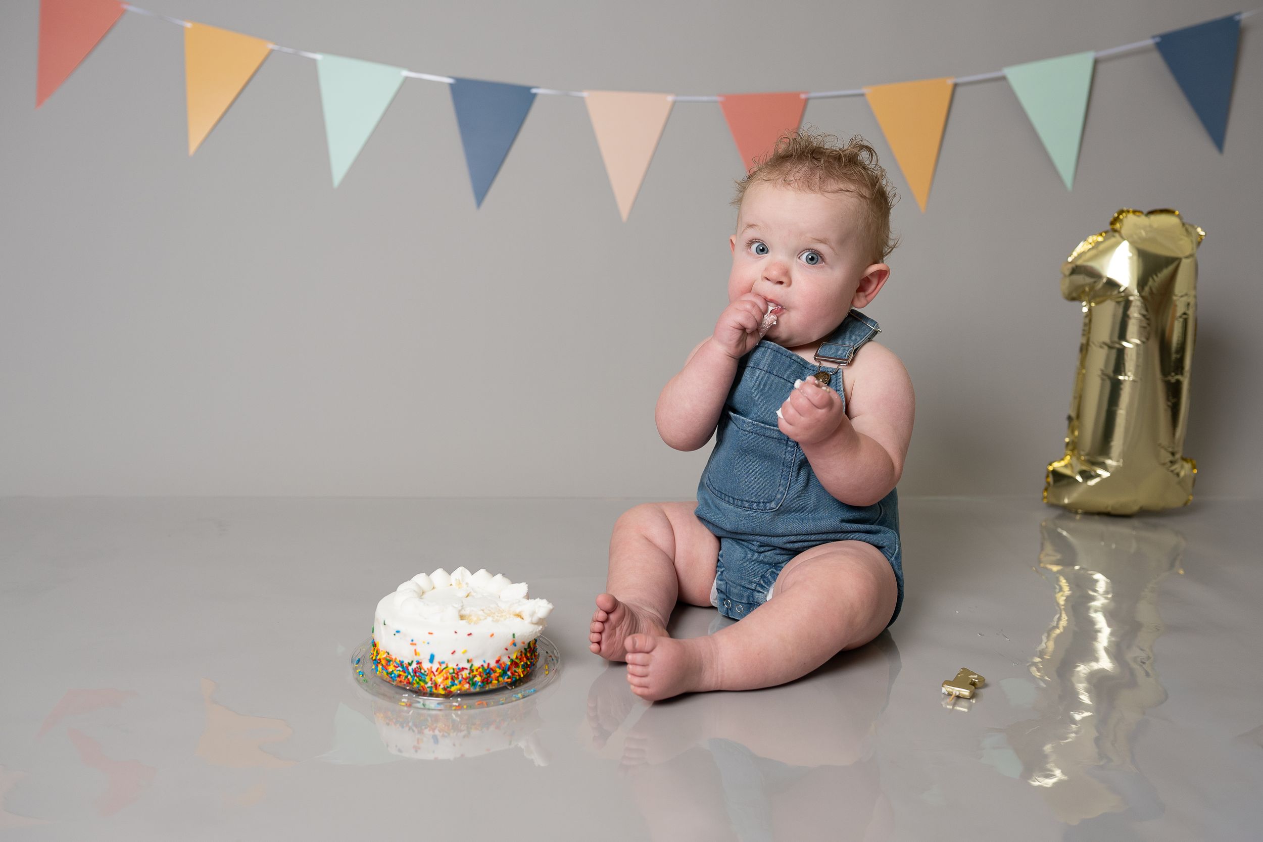 Baby in blue romper eating first birthday cake beside a gold number one balloon and colorful bunting.