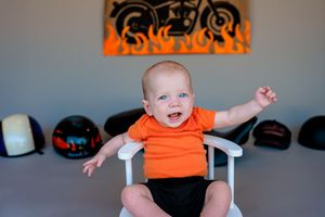 Small infant wearing an orange t-shirt and black shorts sits in a chair with motorcycle helmets displayed on wall behind them.