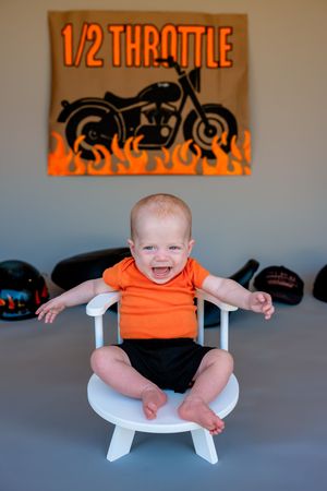 A baby wearing an orange shirt sits in a modern white chair against a gray wall with orange artwork in the background.