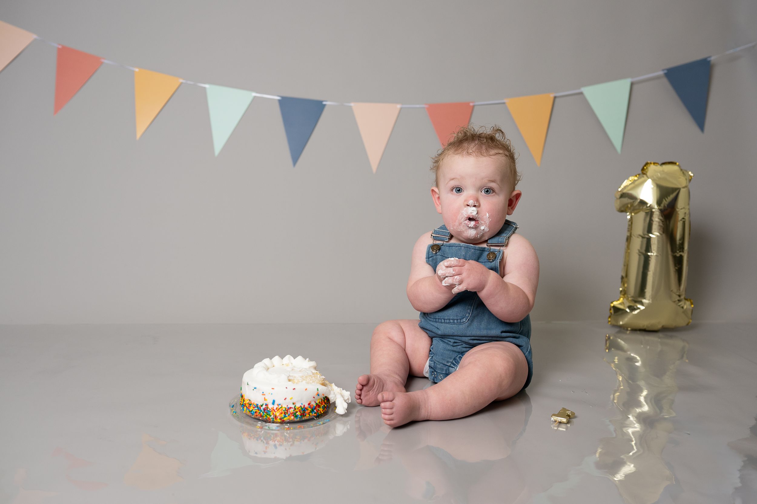 Baby's first birthday cake smash photo with gold number one balloon and colorful bunting decorations.