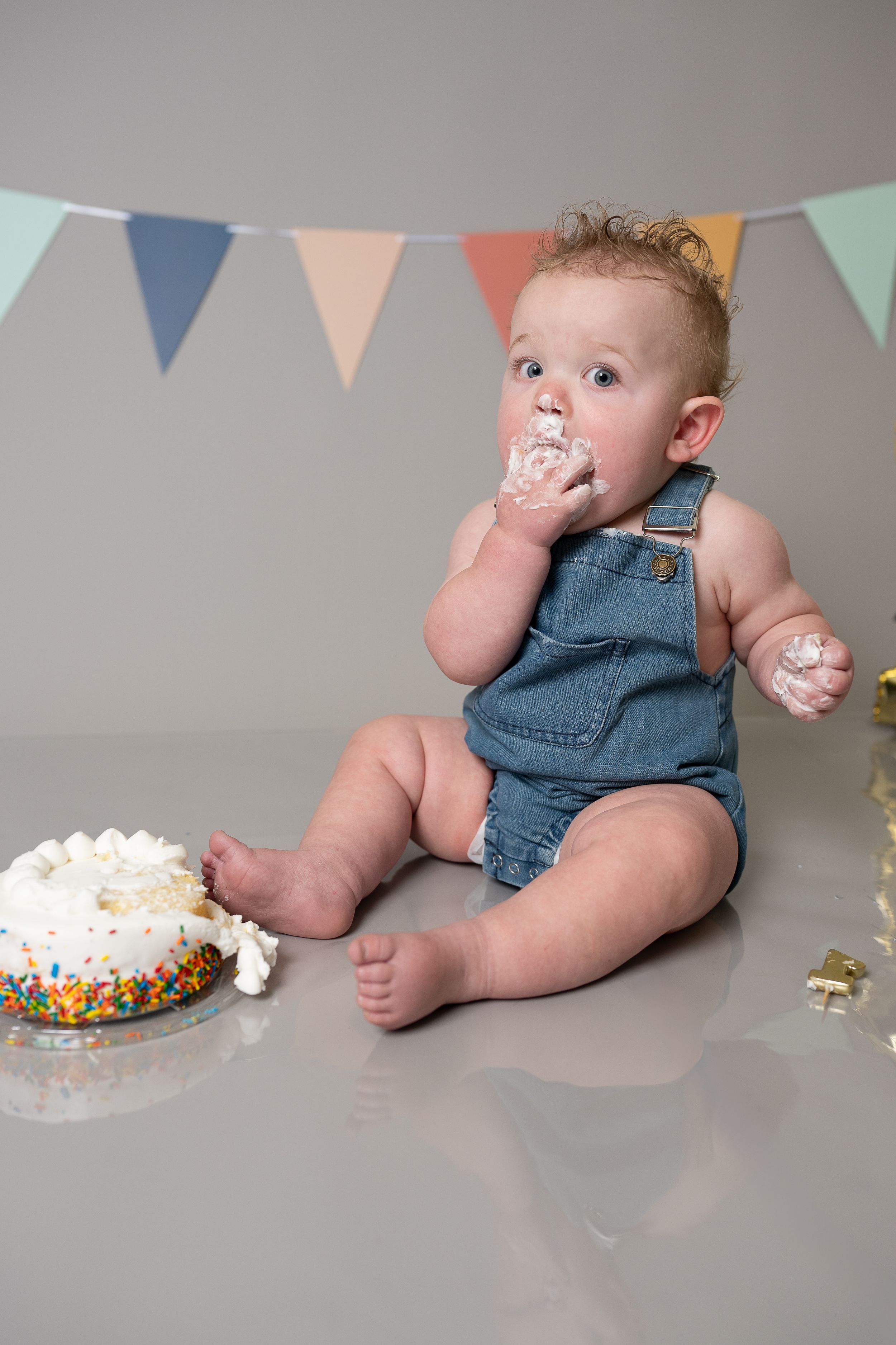 Baby in denim overalls eating first birthday cake with colorful sprinkles and bunting flags in background.