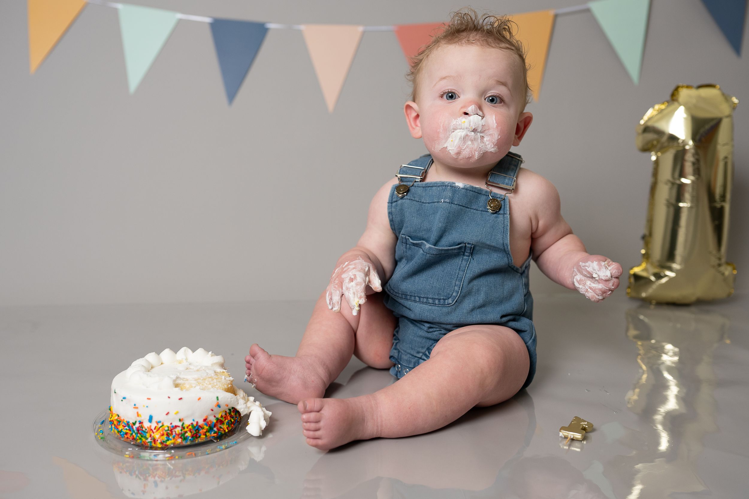 Baby in denim overalls smashing first birthday cake with frosting on face, sitting near gold number one balloon.
