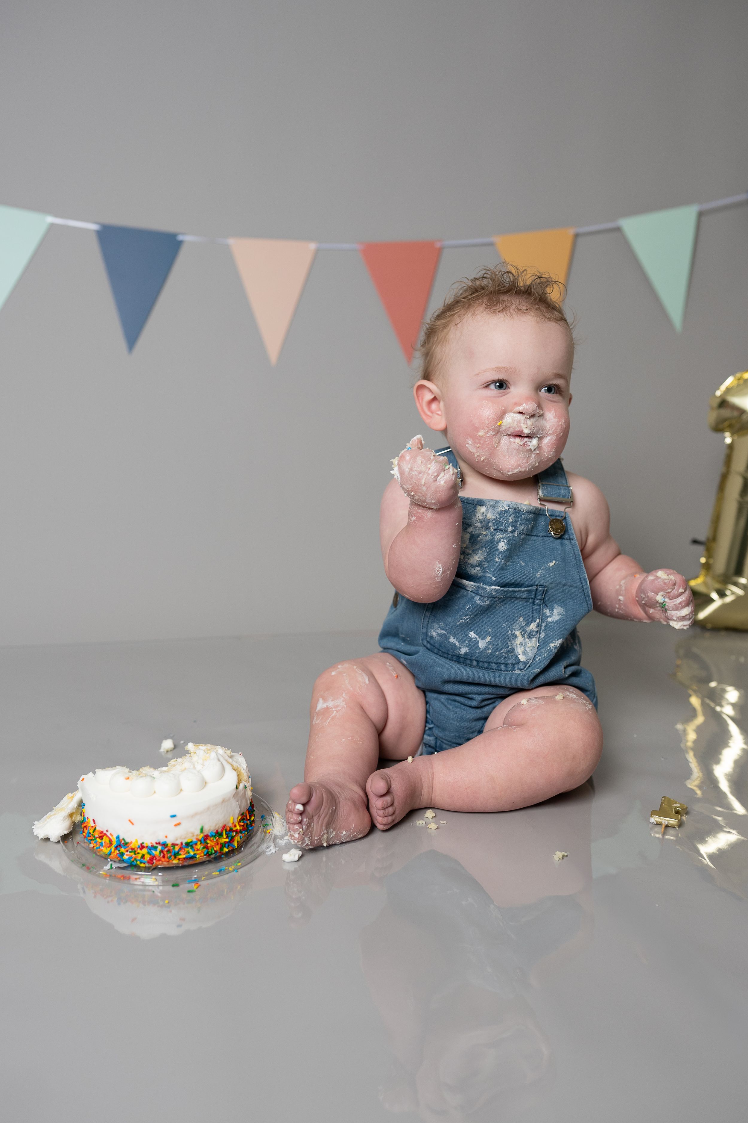 Baby in denim overalls celebrates first birthday smashing cake, covered in frosting, with colorful bunting and gold number one balloon.