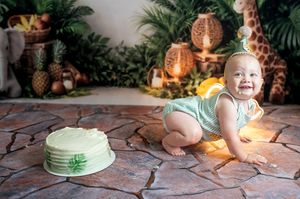 Baby smiling at jungle safari first birthday cake smash with giraffe backdrop and tropical decorations.