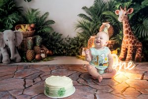 Baby's first birthday cake smash with jungle safari theme, stuffed giraffe and elephant, tropical plants, and glowing 'one' sign.