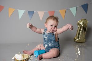 Baby celebrates first birthday smashing cake, wearing denim romper, with colorful bunting and gold number one balloon.
