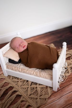Sleeping newborn in brown knit wrap on white vintage-style doll bed with macrame rug beneath.