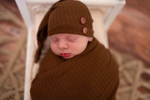 Sleeping newborn baby swaddled in brown knit blanket with matching button beanie on miniature white bed.