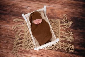 Newborn baby swaddled in brown wrap sleeping peacefully on white miniature bed with rustic wooden floor background.