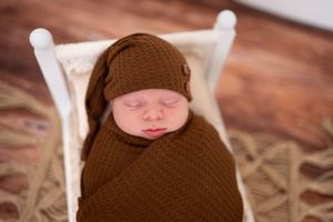 Peacefully sleeping infant wrapped in brown knitted swaddle with matching hat on white wooden prop bed.