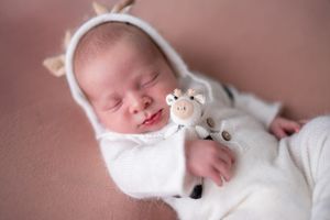 Peaceful newborn baby wearing white swaddle sleeps on dusty rose colored backdrop in professional photo style.