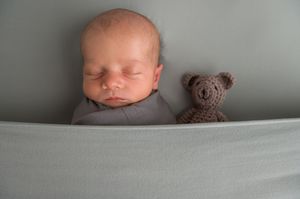 Sleeping newborn laid on light gray backdrop with small brown teddy bear nearby.
