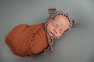 Sleeping newborn wrapped in a rust colored swaddle and wearing a knit bonnet.