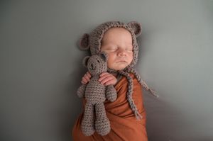 Baby wearing knitted bear hat and orange swaddle resting peacefully on gray backdrop.