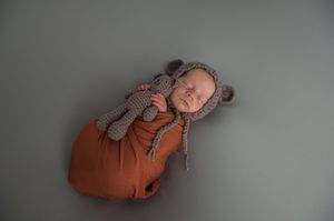 A sleeping infant lies peacefully on a gray backdrop wearing a pumpkin-colored outfit with a knit stuffed toy.