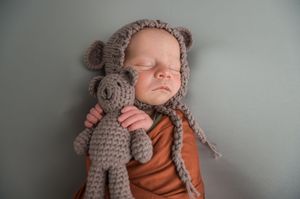 A newborn baby rests on a gray background wearing orange clothing while snuggled with a crocheted stuffed animal.
