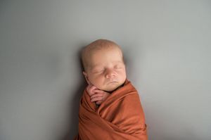Newborn baby wrapped in rust-colored swaddle blanket sleeping peacefully against grey background.