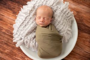 Newborn baby in olive wrap nestled on white textured blanket against wooden backdrop.
