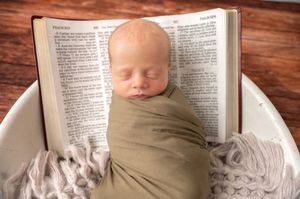 A newborn baby peacefully sleeps on an open Bible on a wooden surface in soft lighting.