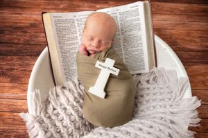 A small baby rests atop an open Bible while holding a white cross on a textured blanket.