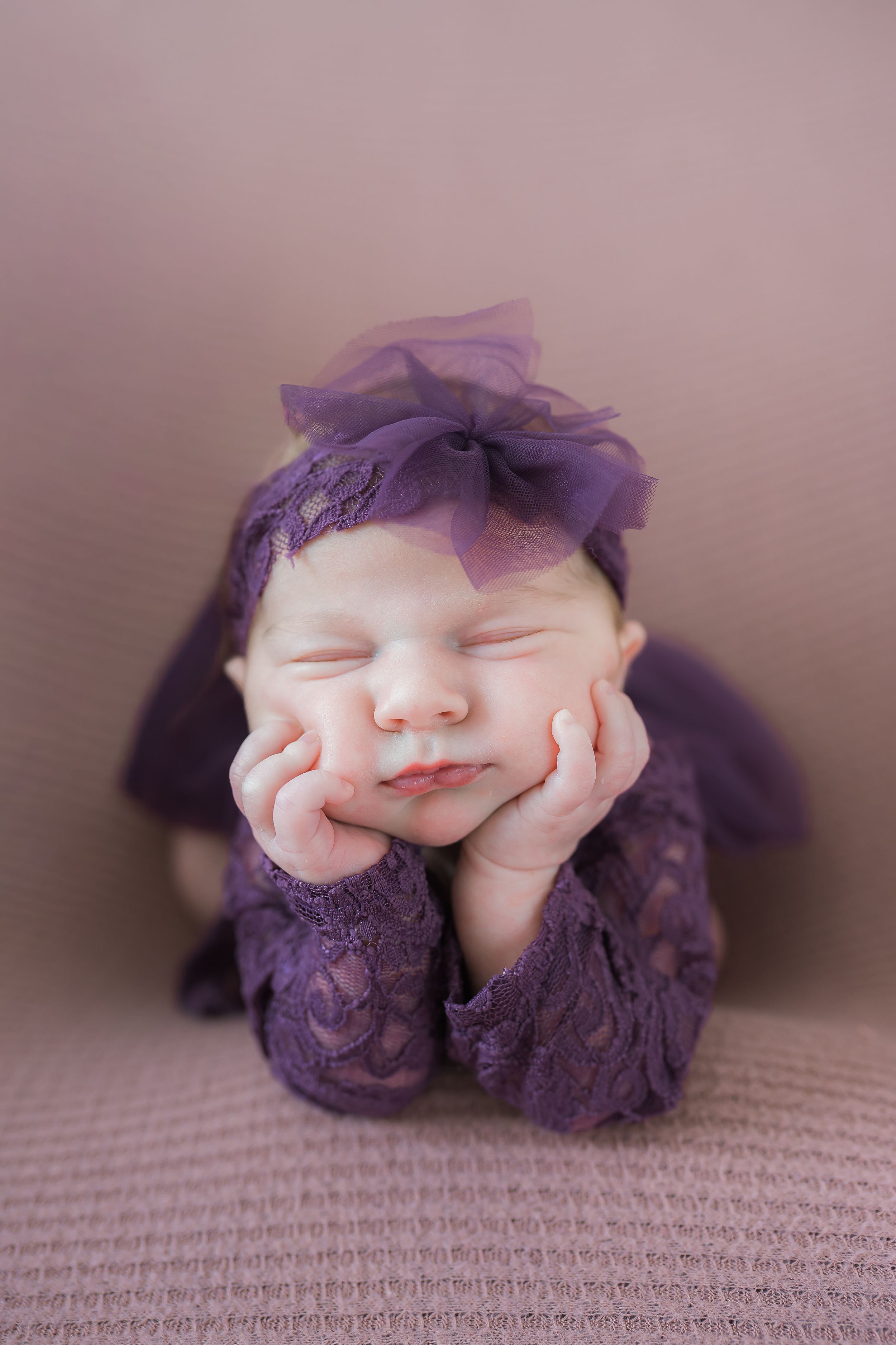 Sleeping newborn resting on textured fabric wearing purple lace outfit and matching headband in soft lighting.
