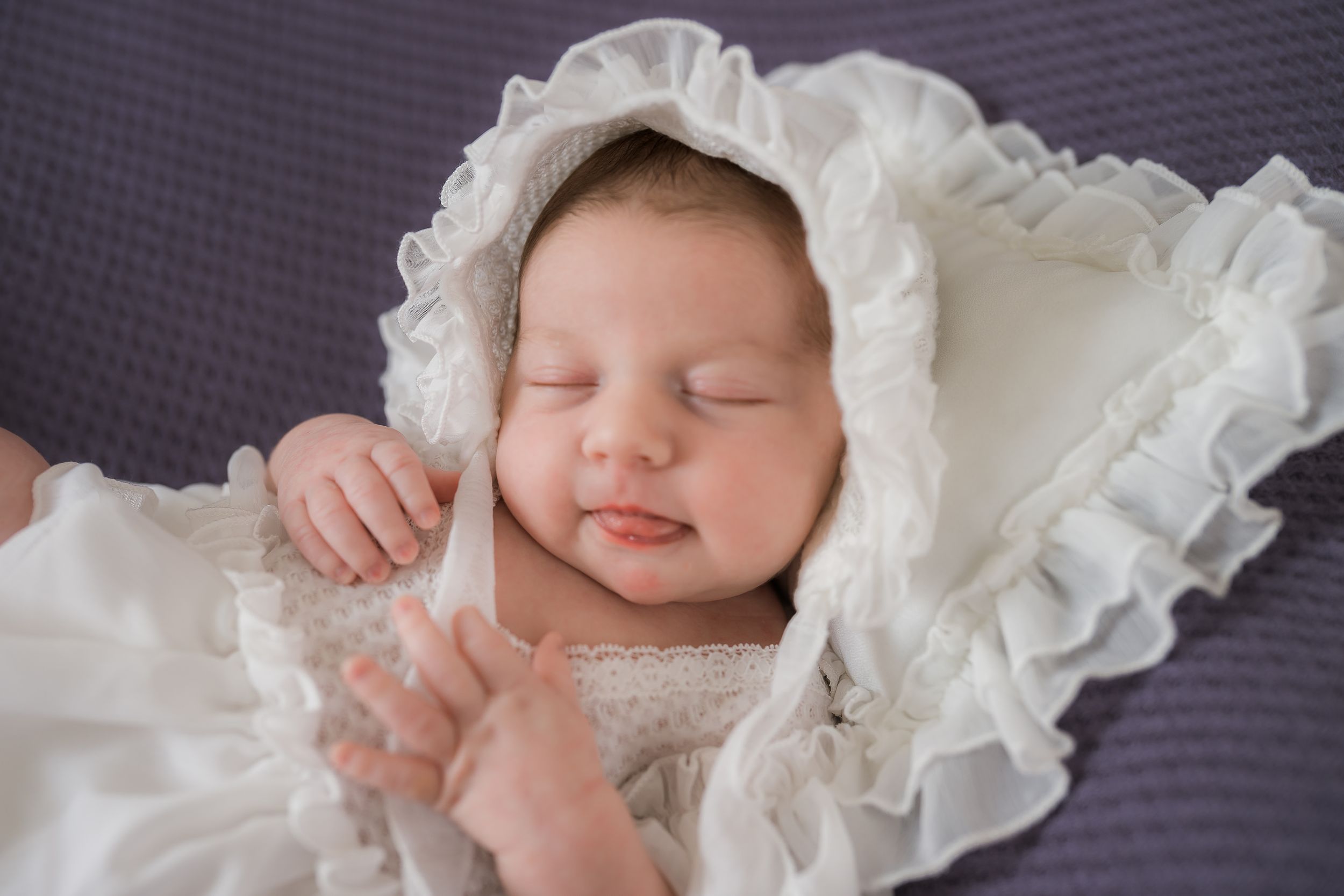 Sleeping newborn in ornate white christening gown and bonnet rests peacefully on gray background.