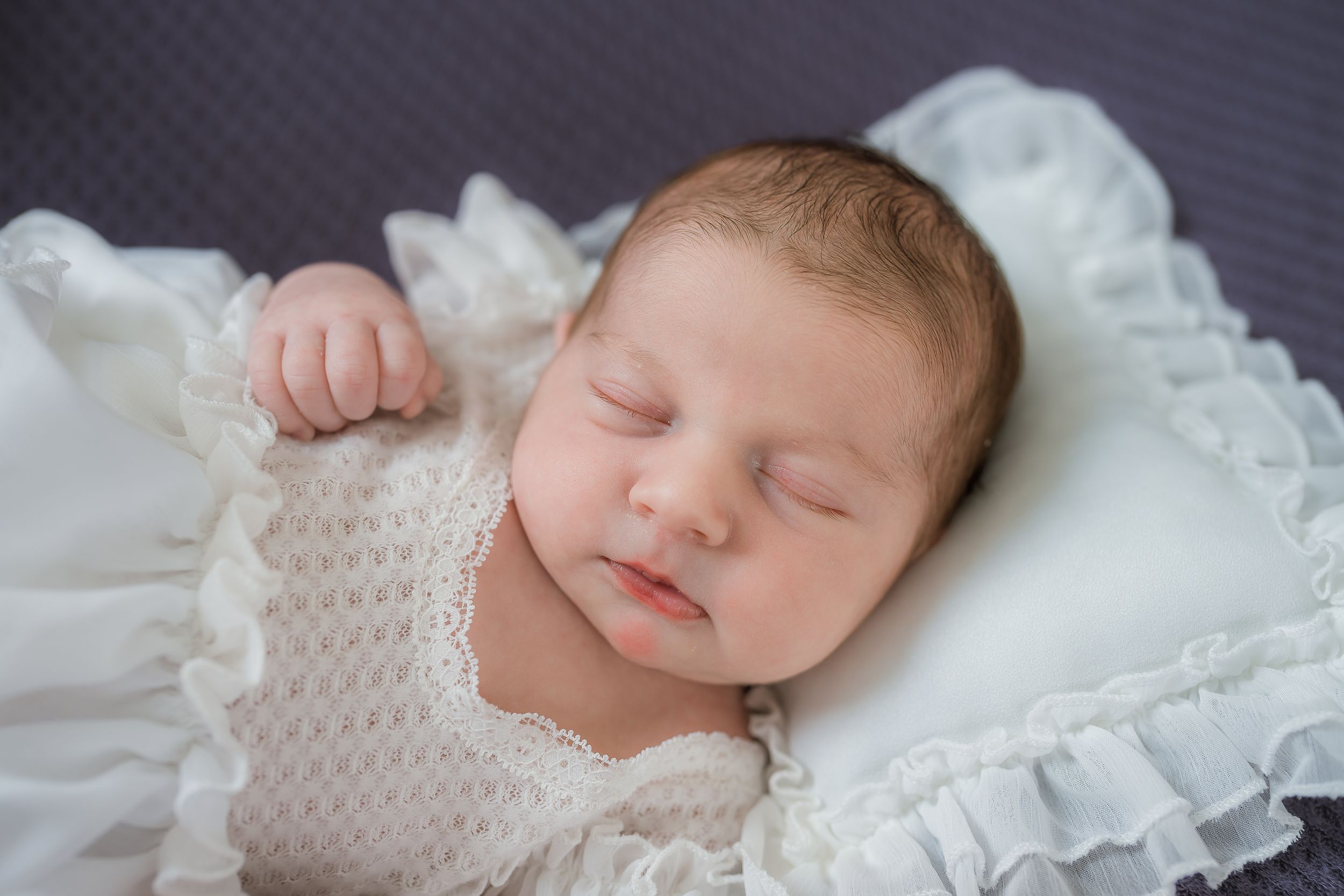 Newborn baby sleeping peacefully on white textured blanket with knitted pillow detail.