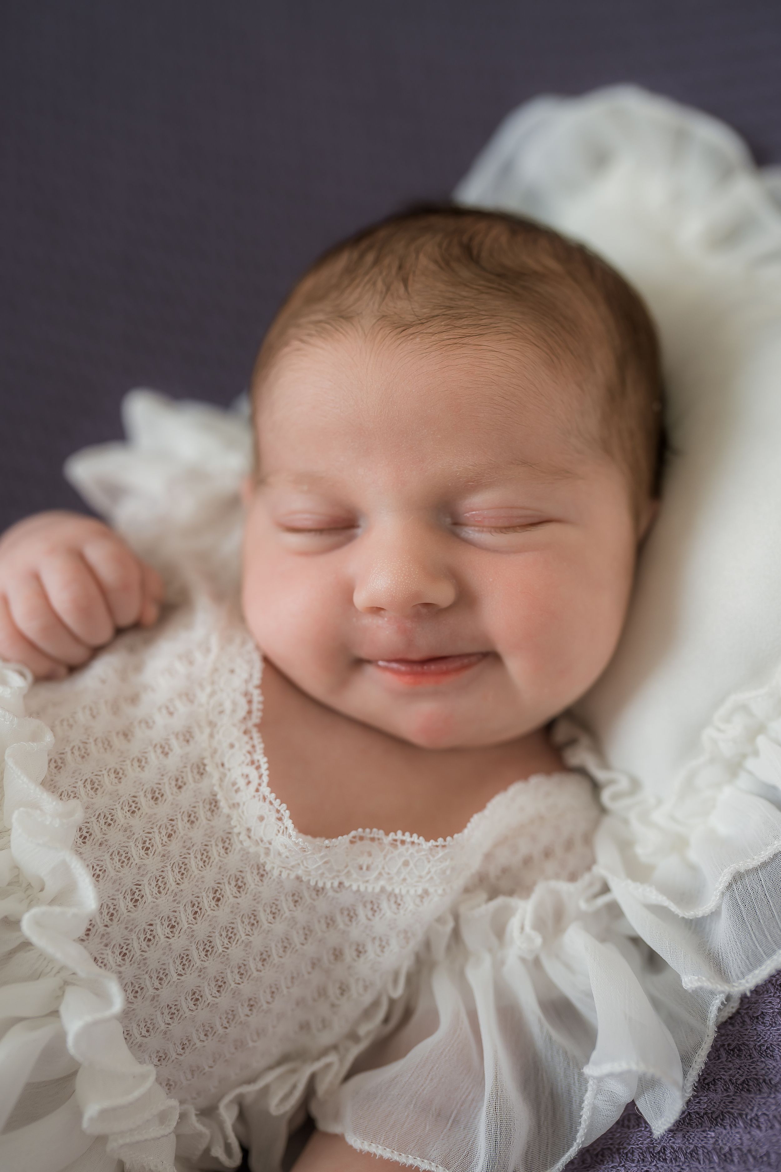 A sleeping newborn baby wearing a white lacy dress smiles peacefully in a series of close-up portraits.