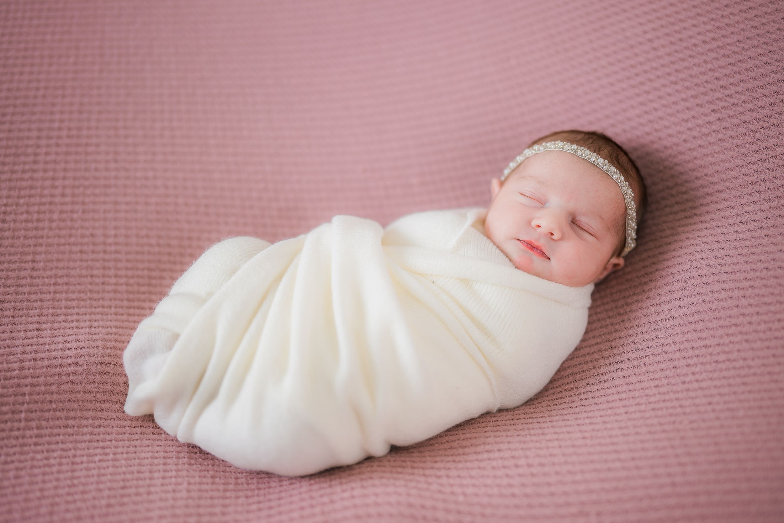 Newborn baby wrapped in white fabric lying on pink textured backdrop during photo session.