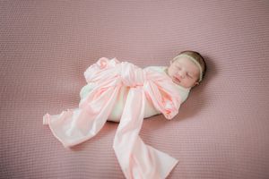 Sweet newborn baby sleeping peacefully while wrapped in delicate pink and white wrap on textured background.