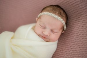 A newborn baby peacefully sleeping while wrapped in a soft white blanket and wearing a delicate cream-colored headband.