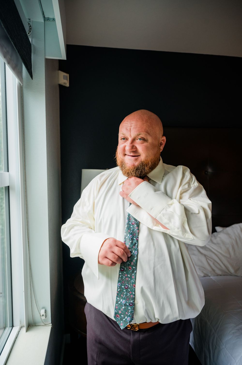 Groom in white dress shirt and patterned tie getting ready in hotel room.
