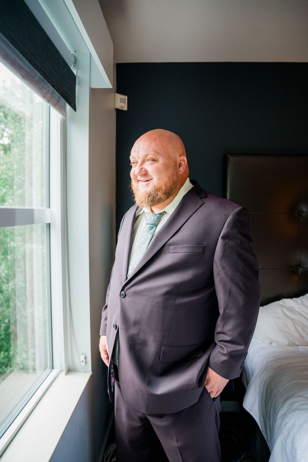 Groom in dark suit poses by window during wedding preparations.