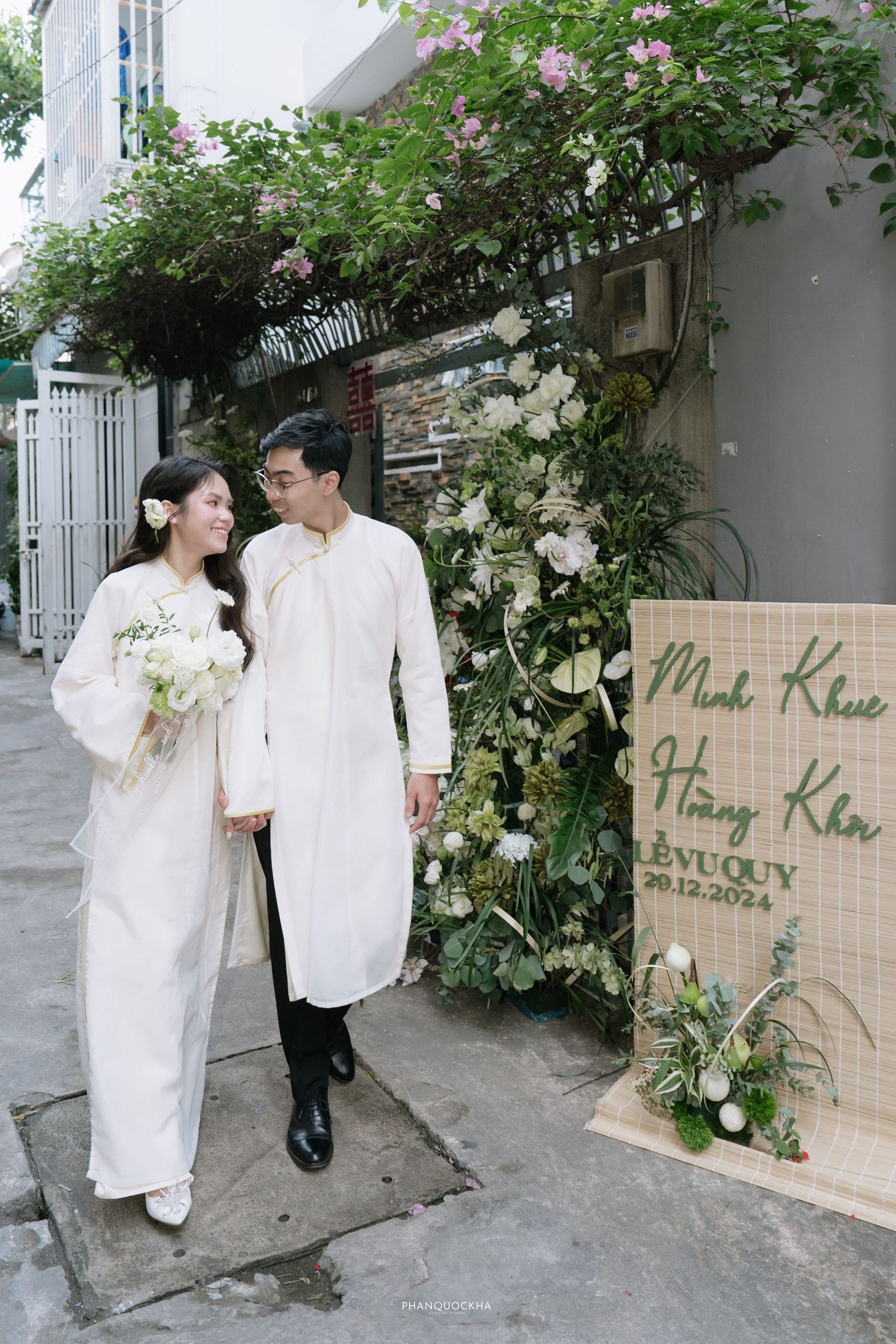 Vietnamese couple in white ao dai walk hand in hand past floral wedding arch and bamboo sign.
