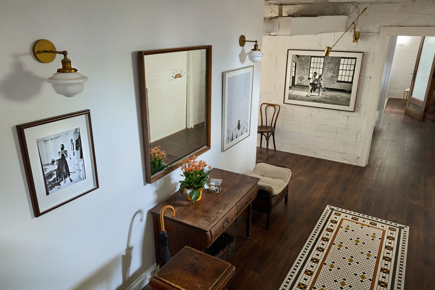 Long hallway with hardwood floors, framed artwork, and vintage furniture along white walls.