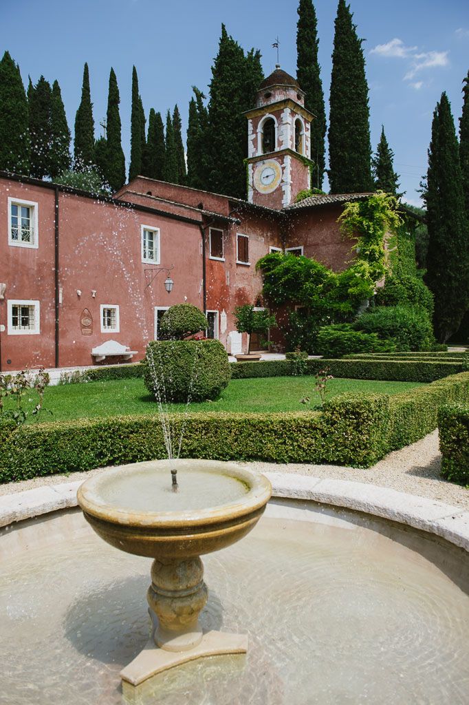 Elegant garden fountain with cypress trees and a pink Mediterranean villa creates a serene Italian countryside scene.