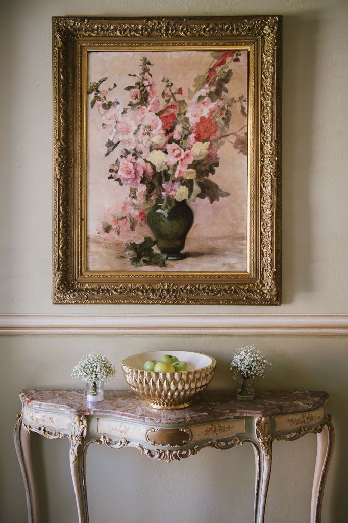 Antique gold-framed floral paintings hang above ornate marble-topped console tables with carved details against a cream wall.