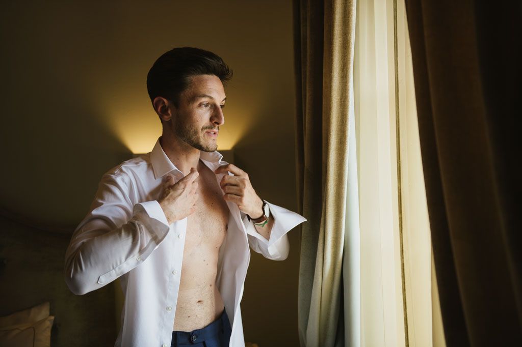 Model poses by window wearing white button-up shirt in dramatic lighting and artistic black and white tones.