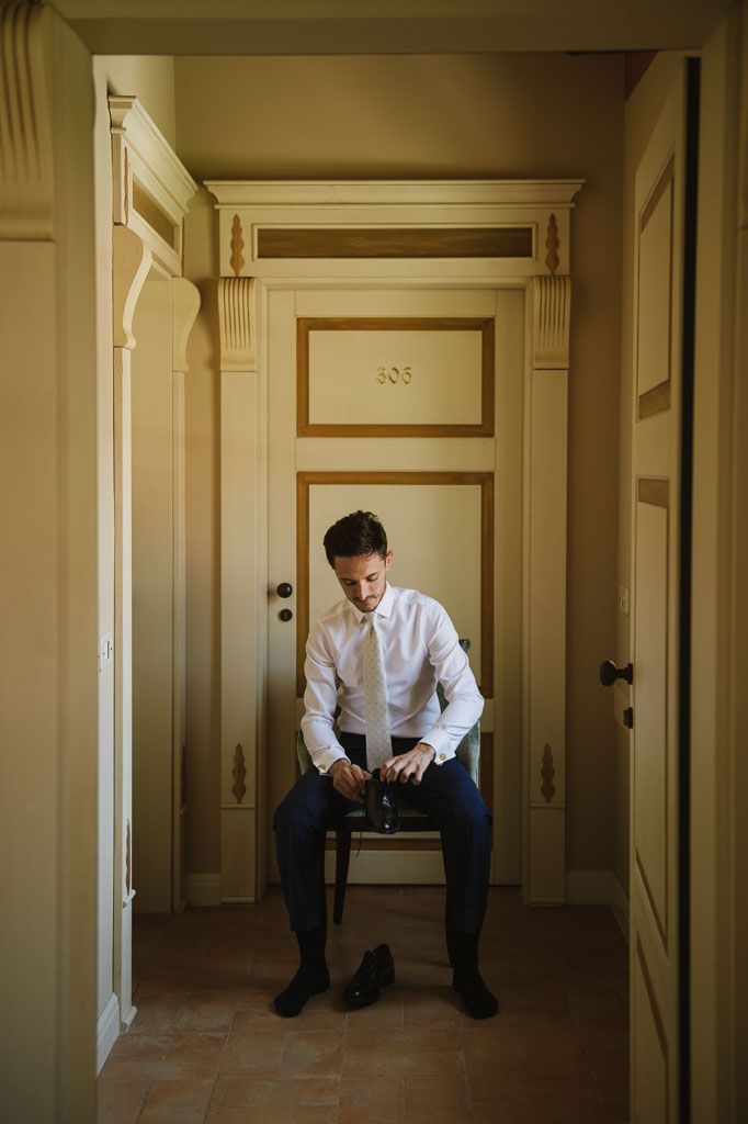 A person in a white shirt and dark pants sits alone in a bare hallway with cream colored walls and doors.