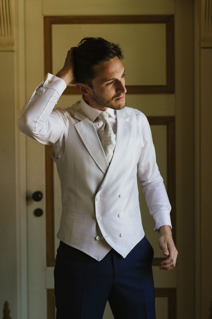A man in a white dress shirt and vest poses against a wall during formal preparations.