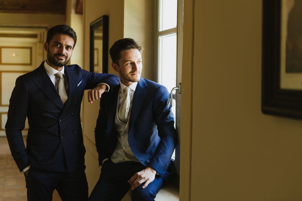 Two people in dark suits share an intimate moment in a warmly lit hallway with windows.
