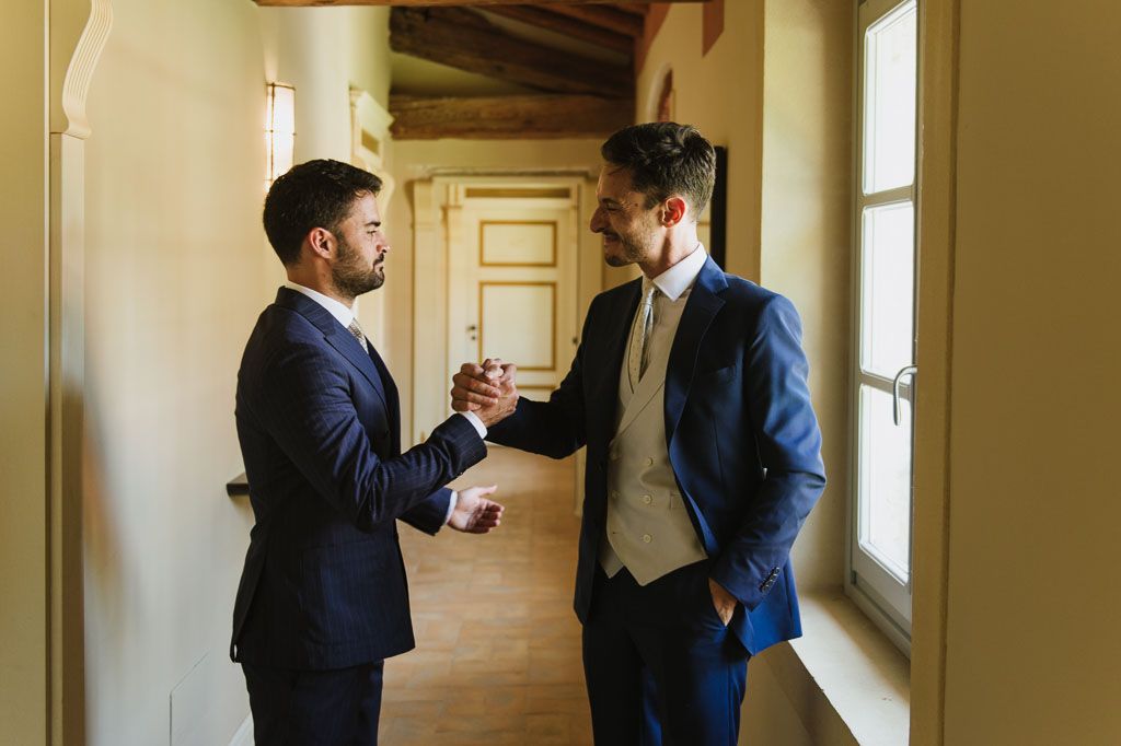 Two people in formal attire having a candid conversation in an ornate hallway with natural lighting.