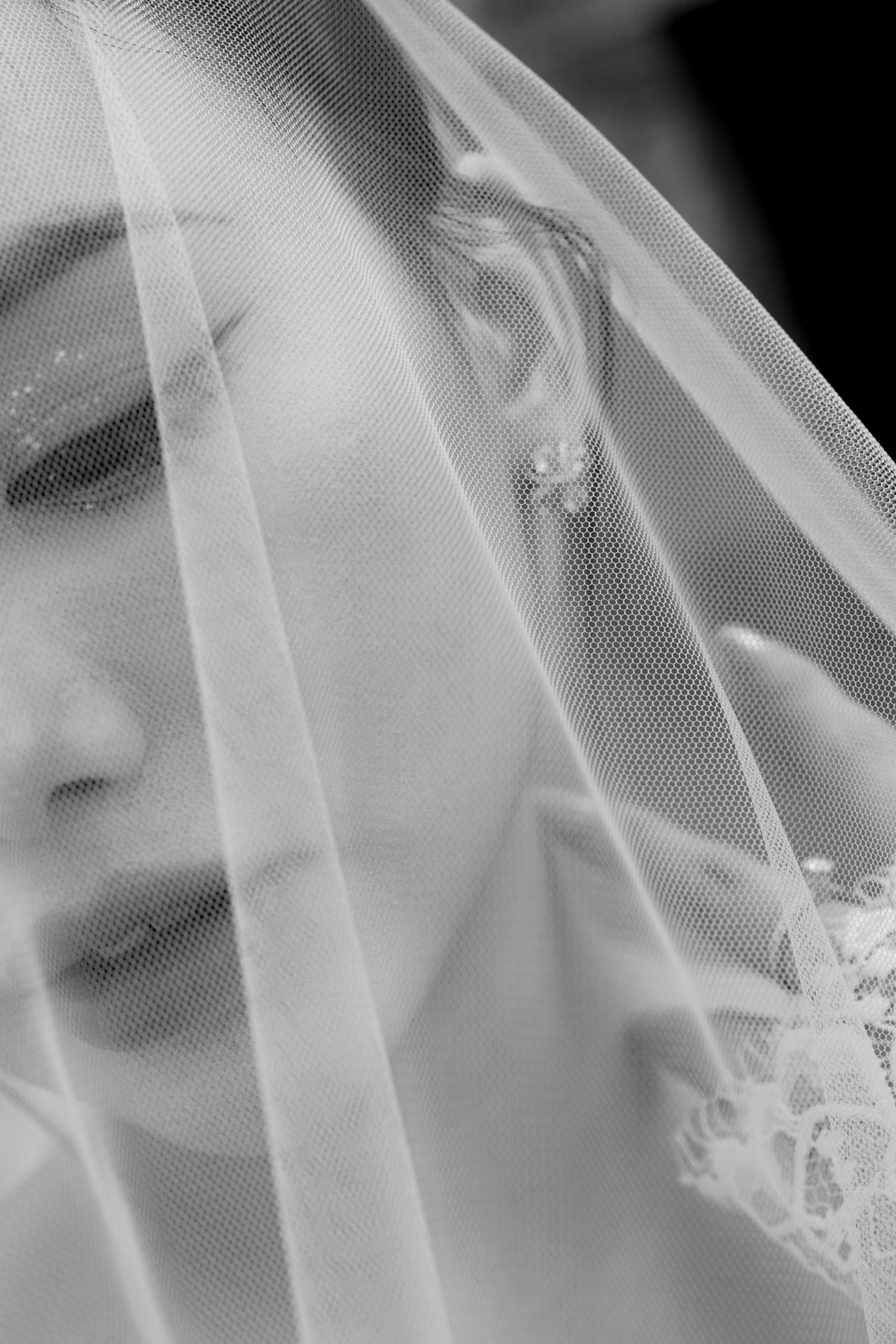Intimate black and white close-up of a bride with a sheer lace veil covering her face, wearing a floral earring.