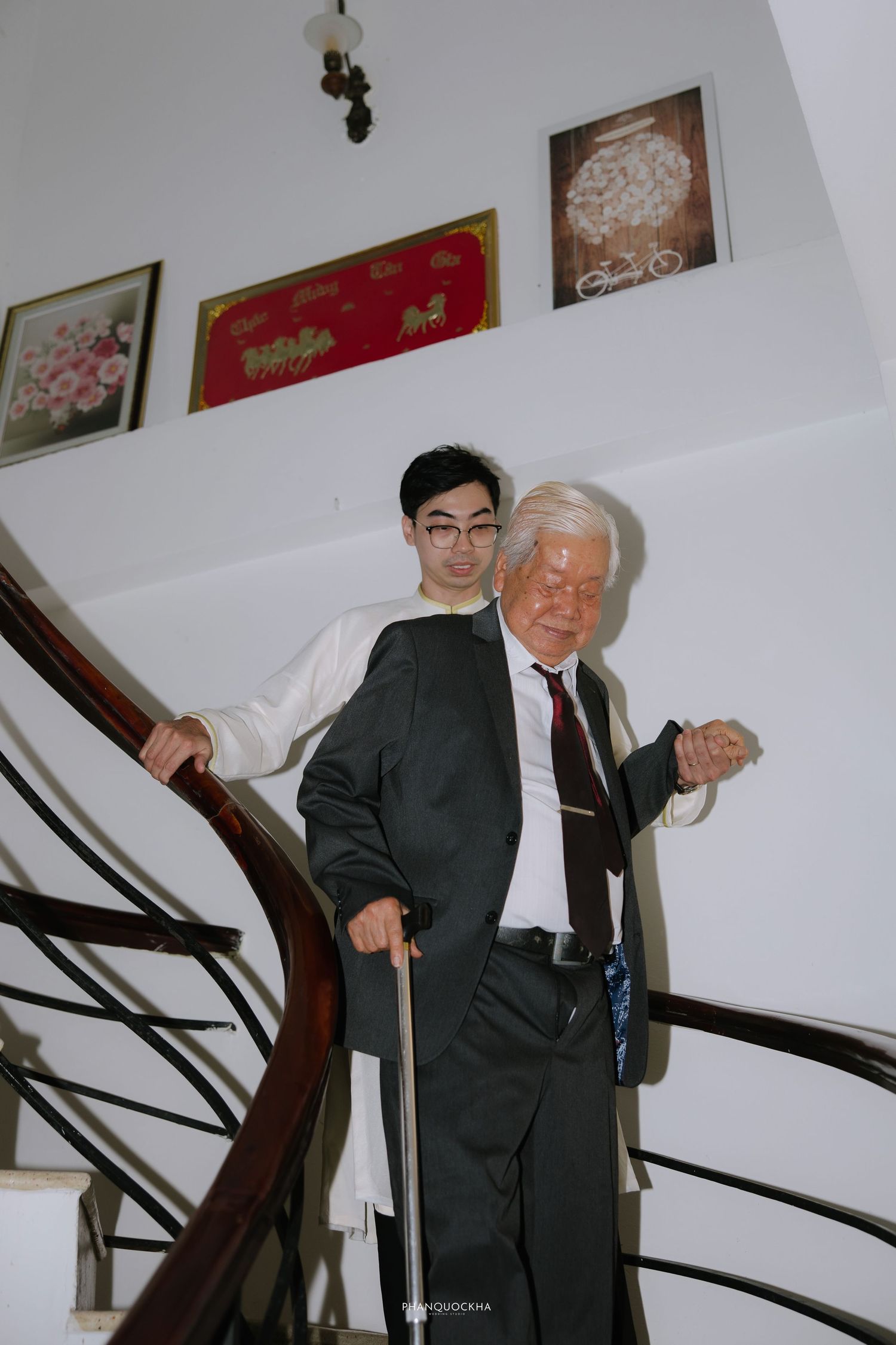 Young Vietnamese groom in white ao dai assists elderly man down wooden staircase indoors.