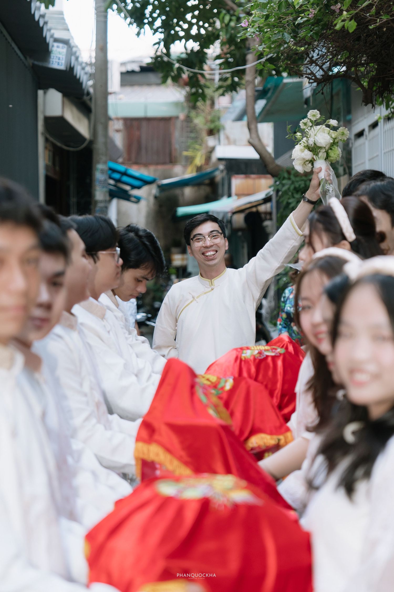 Smiling groom in white ao dai raises bouquet while leading wedding procession down narrow alley.