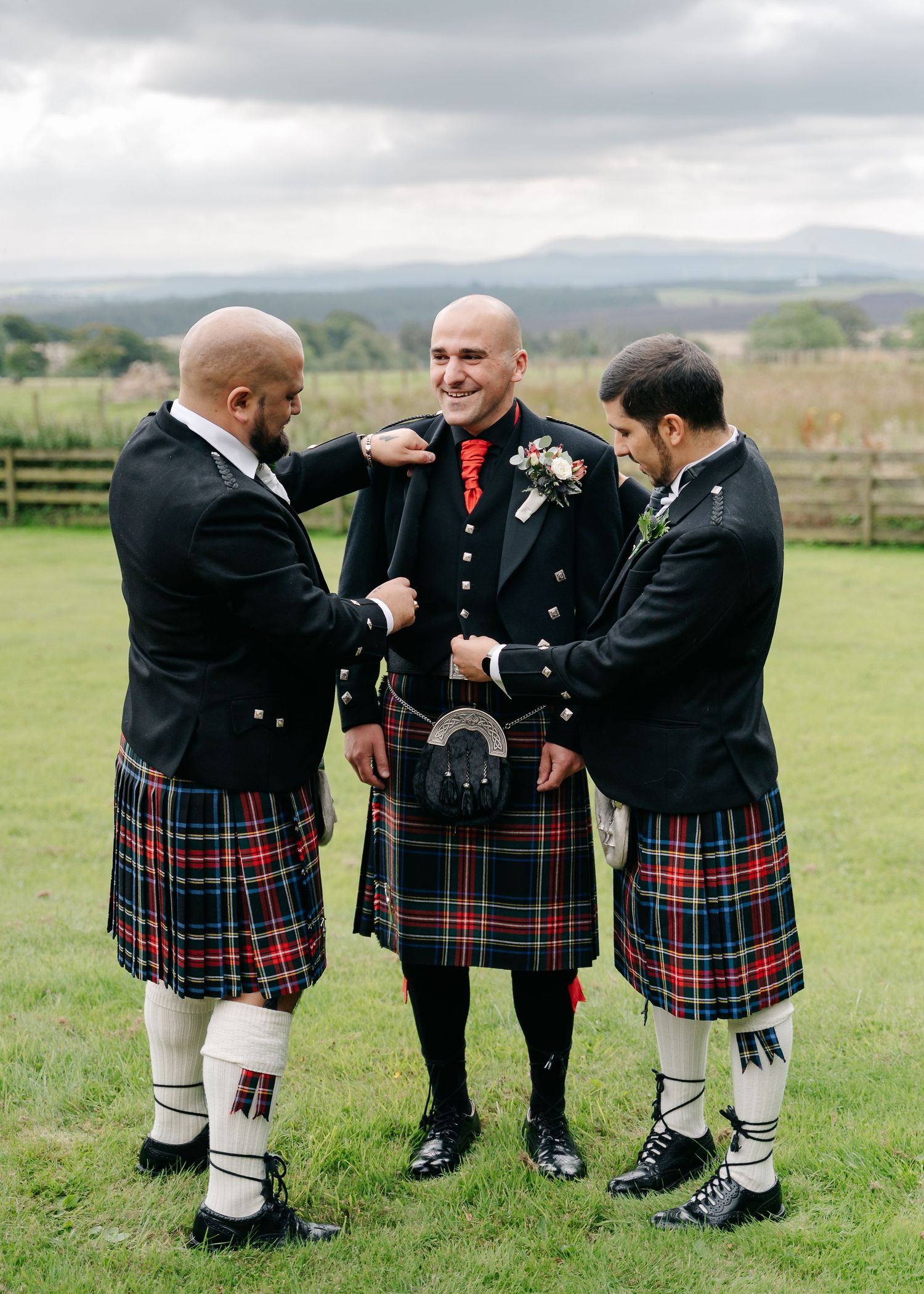Three men in traditional Scottish kilts and formal wear sharing a moment on grassy hillside.