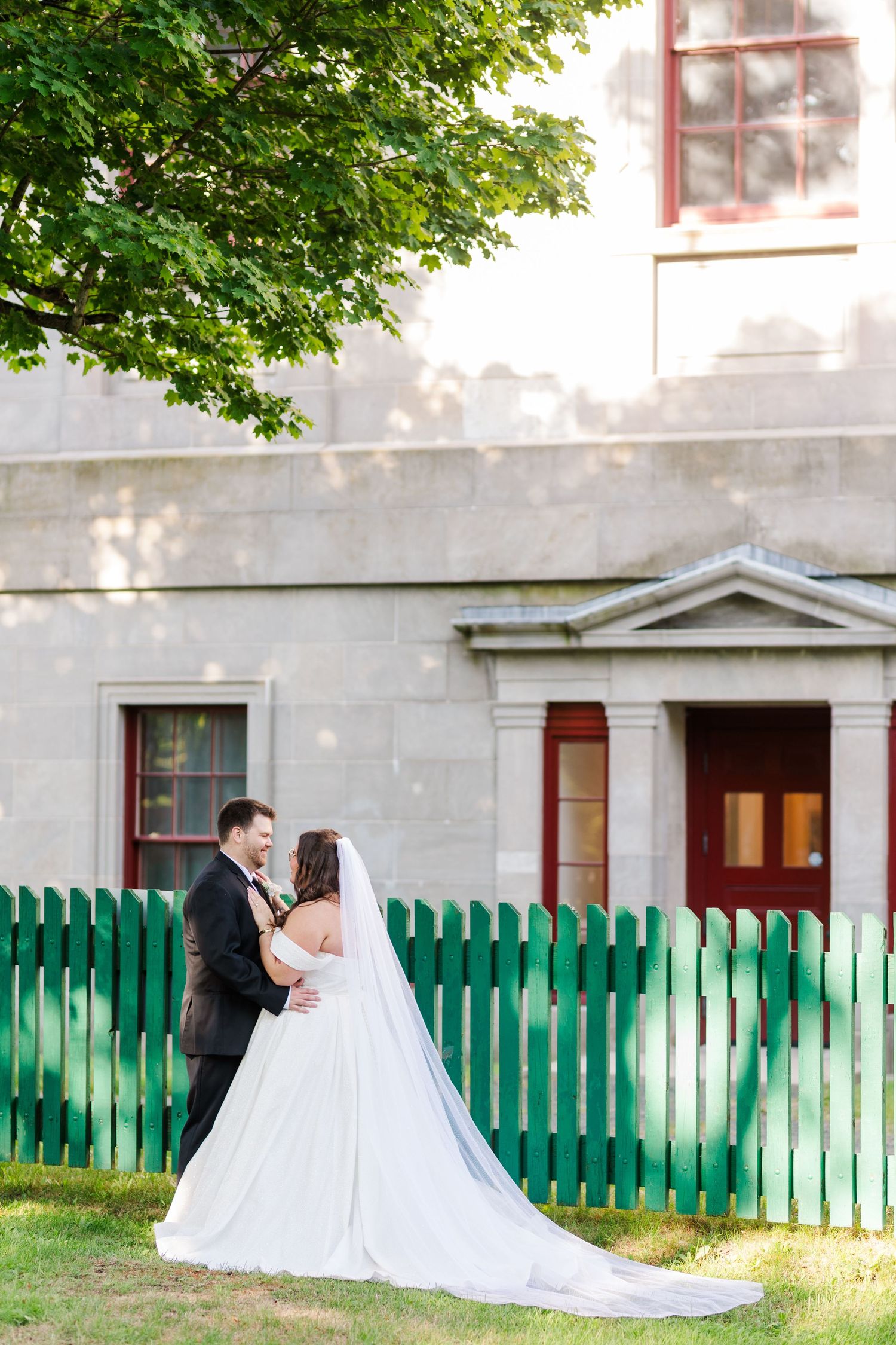 A couple in wedding attire embrace by a white building with a green picket fence on a sunny day.